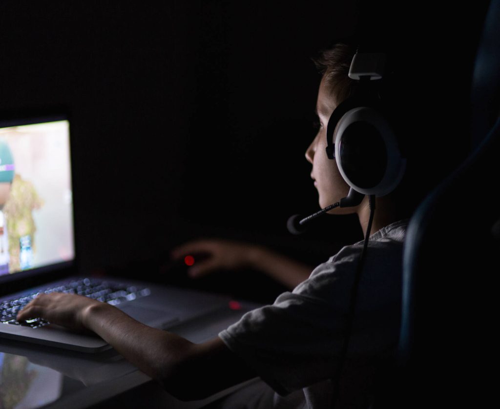 A young child sits at a laptop in the dark with headset on