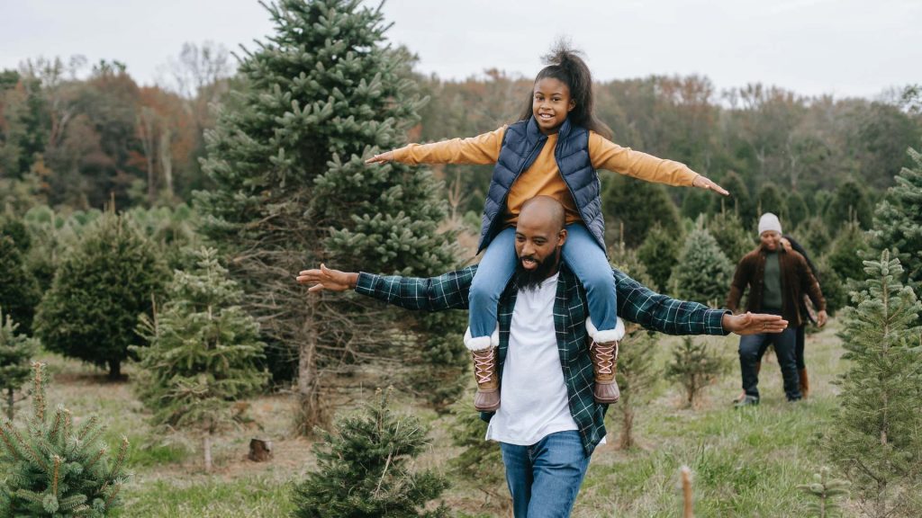 a father is walking in a green forest area with arms outstretched giving his daughter a piggy back. Her arms also outstretched.