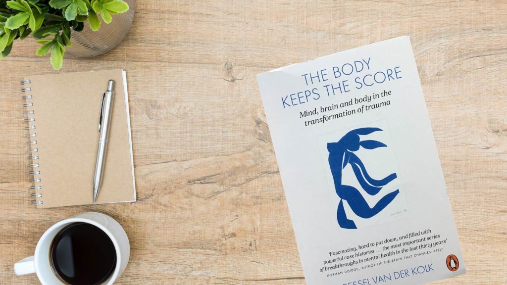 The Body Keeps The Score (book) on a desk alongside a notepad, coffee and a plant
