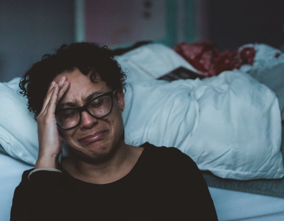 someone with short curly hair and glasses crying with their hand on their face beside a bed.