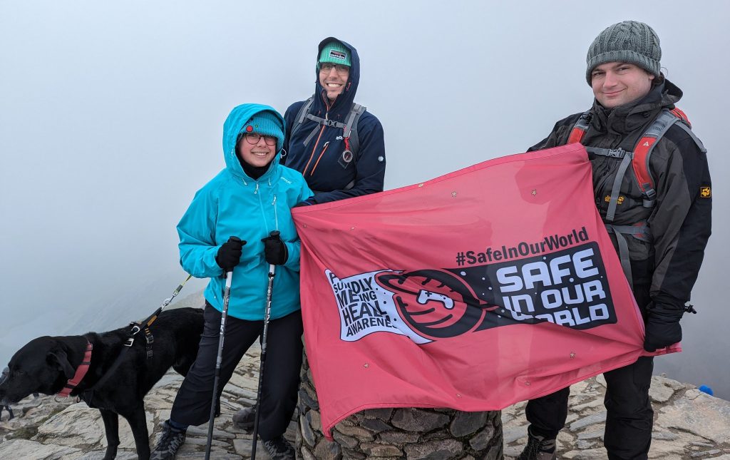 Em, Communications and Events Manager at Safe In Our World, at the summit of Snowdon, Wales, with two friends, holding a Safe In Our World charity flag. Image attributed to article about absence anxiety.