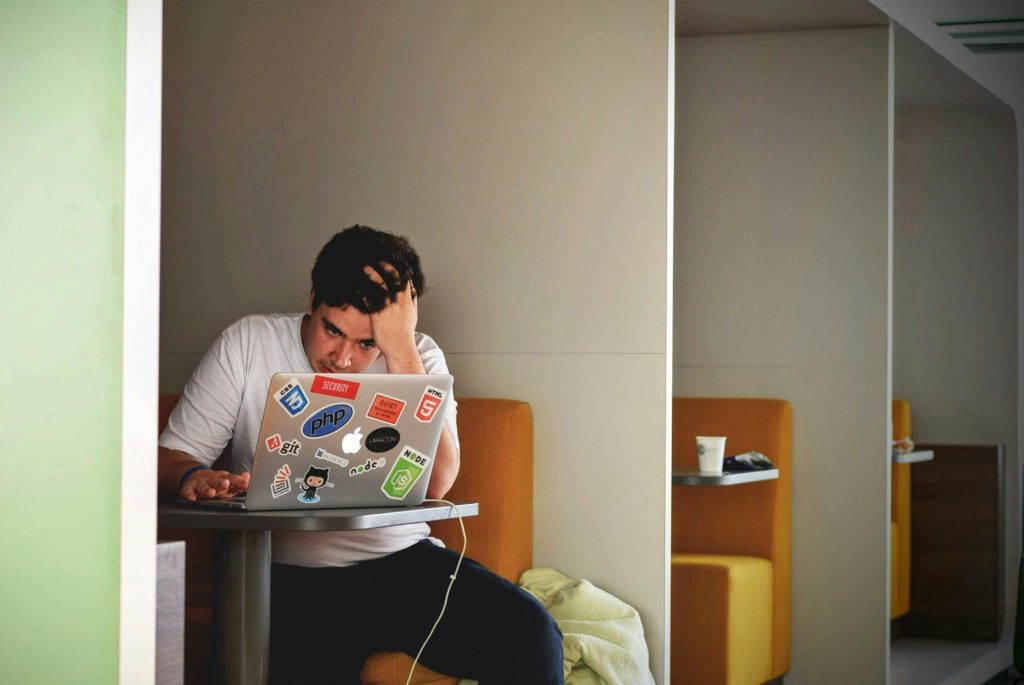 A person sits in a booth at a working cafe, frustratedly holding their head while looking at a laptop screen.