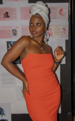 Msani Riann, wearing an orange dress and hoop earrings, looking off to the side and posing for a photo.