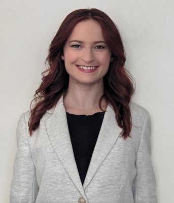 A photo of Maura, a woman with long reddish brown hair, wearing a black top and light grey suit jacket, smiling.