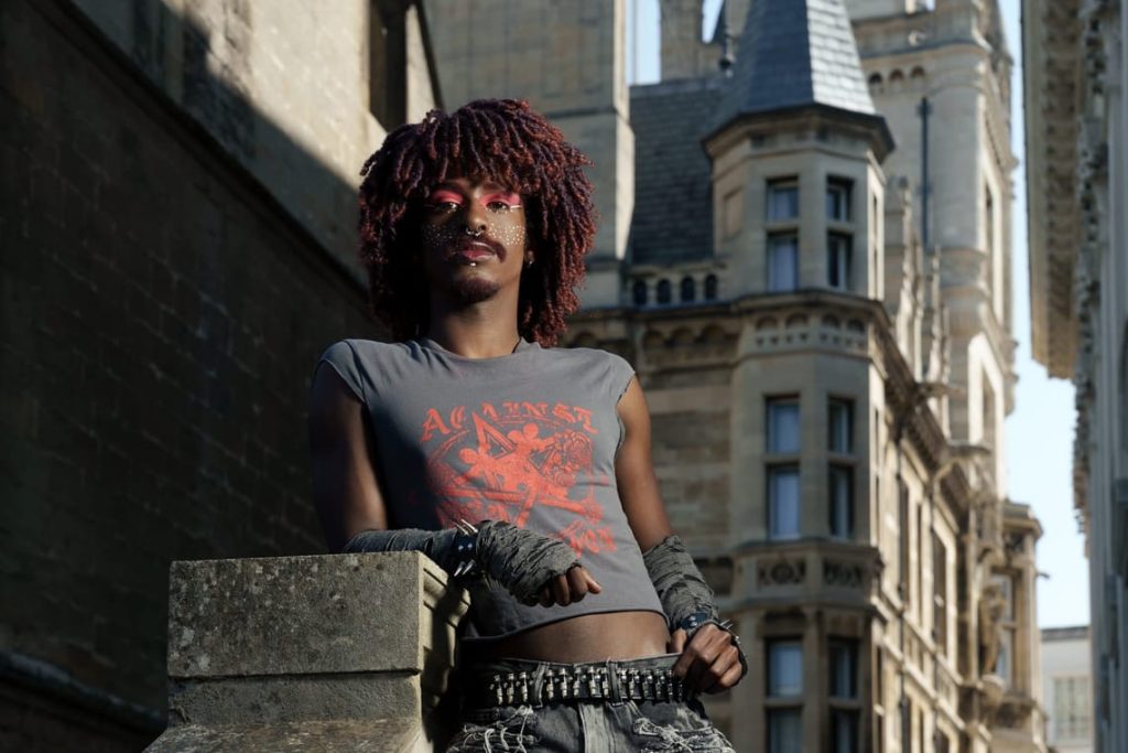 A photo showing drag performer, Guillotina, with curly brown hair tinted with red, pink and white makeup against dark skin, and a casual crop t-shirt, fingerless gloves and jeans combo.